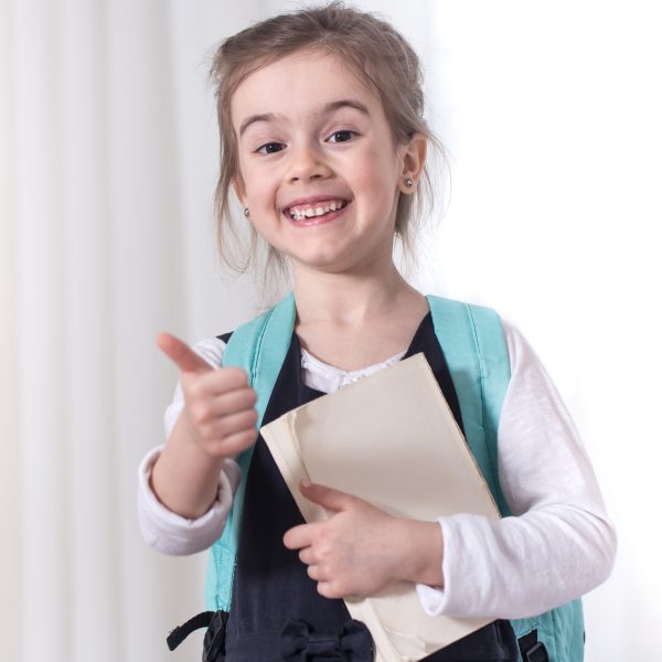Girl-elementary school student with a backpack and a book on a light background .The concept of education and primary school